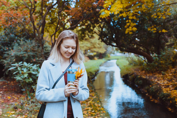 Smiling beautiful young woman in coat enjoying hot drink in her reusable thermos cup and walking in the autumn park near the river. Romantic fall mood. Vintage toning. Selective focus. Copy space. - Фото, зображення
