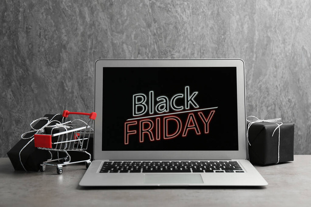 Laptop s oznámením Černý pátek, malý nákupní košík a dárky na stole na šedém pozadí - Fotografie, Obrázek