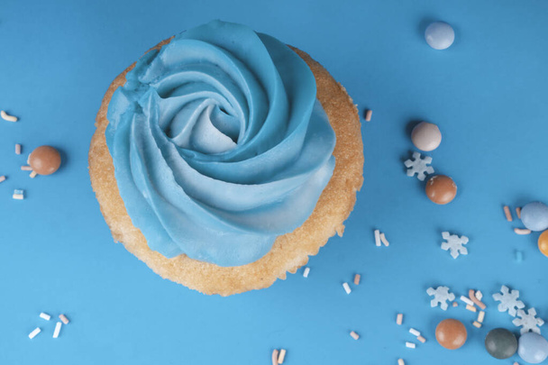 Vanille Cupcake mit Schlagsahne vor blauem Hintergrund, dekoriert mit Zuckerschneeflocken, Streusel, bunten Bonbons, Urlaubskonzept, Grußkarte - Foto, Bild