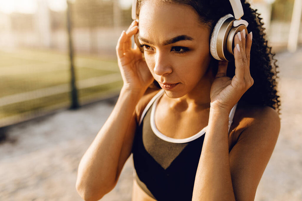 Athletic young african american woman listening to music in headphones, in sportswear outdoors in the park, healthy lifestyle and sport concept - Foto, imagen
