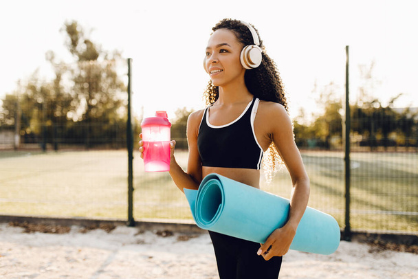 Beautiful young athletic african american woman in sportswear holding a yoga mat and a bottle of water outdoors in the park at sunrise - Foto, imagen