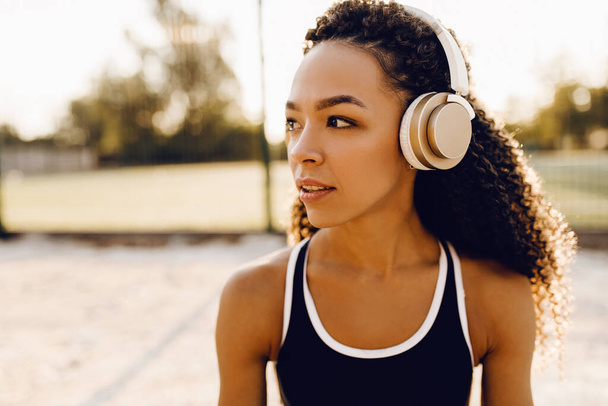 Athletic young african american woman listening to music in headphones, in sportswear outdoors in the park, healthy lifestyle and sport concept - Фото, зображення