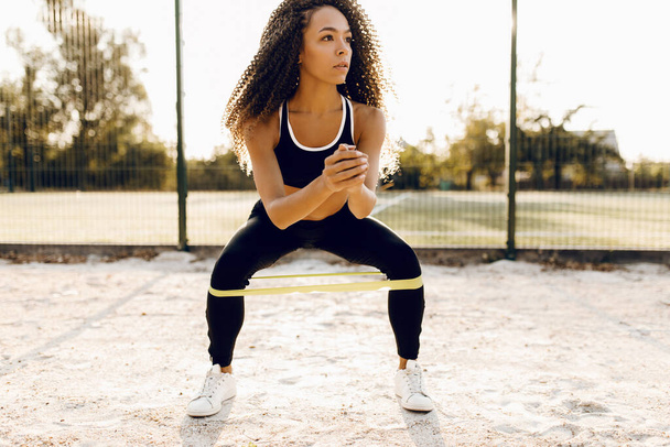 young sportive woman in sportswear, squats with fitness rubber bands, outdoors in the park, sports, healthy lifestyle - Photo, image