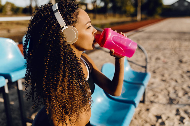 Young athletic african american woman wearing headphones resting after intense jogging and drinking water from a bottle during a break after jogging outdoors - Photo, image