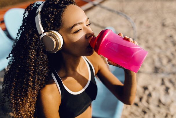 Young athletic african american woman wearing headphones resting after intense jogging and drinking water from a bottle during a break after jogging outdoors - Foto, Bild