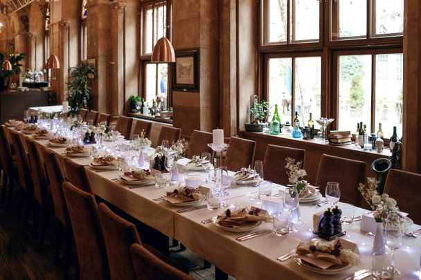 Довгий, покладений стіл в ресторані з безліччю прозорих окулярів, серветок і прикрас для посуду. Глома, темний інтер'єр ресторану в стилі середньовічної фортеці і довгі білі свічки
 - Фото, зображення