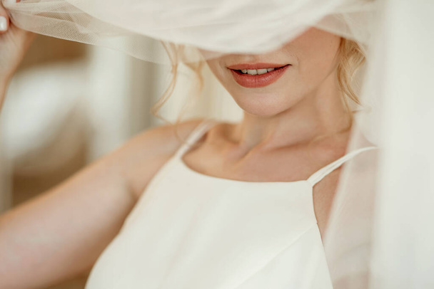 Piękna blond panna młoda w białej sukni ślubnej zakrywa twarz welonem. Skup się na ustach młodej kobiety z czerwoną szminką. Idealna koncepcja narzeczonej - Zdjęcie, obraz