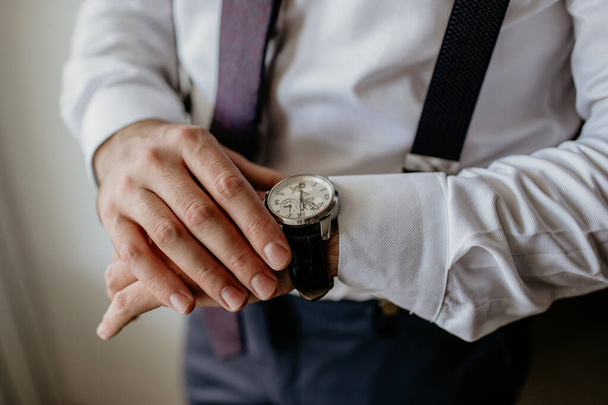 Liikemies klassisissa vaatteissa katsoo kelloa ja hallitsee aikaa. Suspenders ja valkoinen paita ovat osa tyyliä. Mies tarkistaa ajan, ettei myöhästy liiketapaamisesta. - Valokuva, kuva