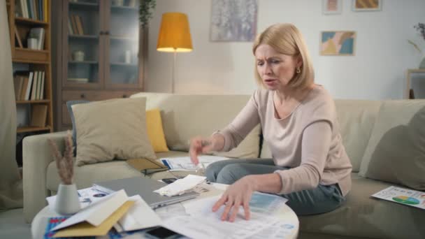 Gestresste Frau mittleren Alters blickt durch Stapel von Papieren auf chaotischem Tisch, während sie die Finanzen analysiert und sich aufregt und frustriert ist - Filmmaterial, Video