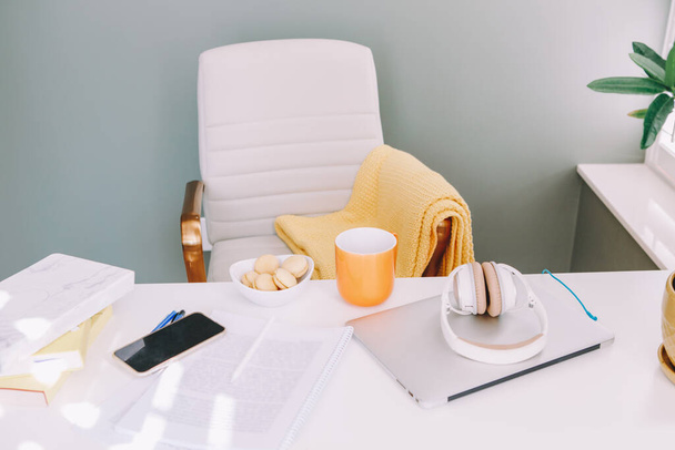 Home office. Cosy comfortable workplace with laptop, headphones, mug, macaroons, smartphone on desk table. Woollen warm blanket on office chair. Work from home. Apartment interior. - Foto, Imagen