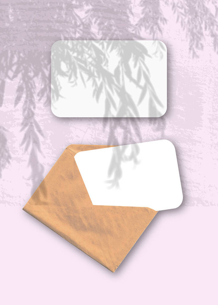 Ein Umschlag mit zwei Blättern strukturiertem weißen Papier auf dem rosafarbenen Hintergrund des Tisches. Mockup-Overlay mit den Schatten der Pflanze. Natürliches Licht wirft Schatten von einem Weidenast - Foto, Bild