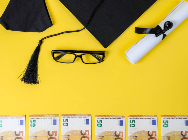 中央のテキストのための場所と黄色の背景に正方形の学術キャップ、卒業証書、眼鏡とお金、トップビューのクローズアップ. - 写真・画像