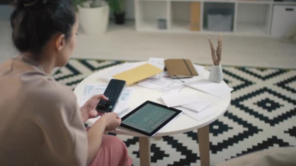 Back-view beelden van jonge gemengde raced vrouw met behulp van calculator app op haar telefoon, terwijl het analyseren van de financiën op tablet zitten in de lichte woonkamer - Video