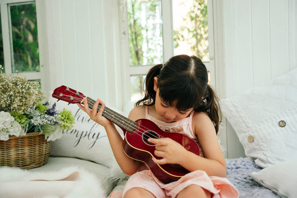 Asian child girl with wooden classic ukulele on sofa at home. - Photo, Image