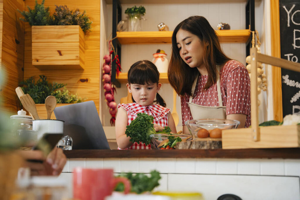 Azjatyckie dziecko i matka pomagające sobie nawzajem gotować w kuchni. Ludzie szukający przepisu na jedzenie w internecie. - Zdjęcie, obraz
