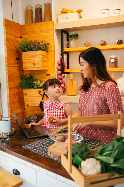 Azjatyckie dziecko i matka pomagające sobie nawzajem gotować w kuchni. Ludzie szukający przepisu na jedzenie w internecie. - Zdjęcie, obraz