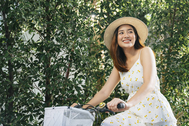 Beautiful asian woman wear hat and white dress riding bicycle at park. - Zdjęcie, obraz