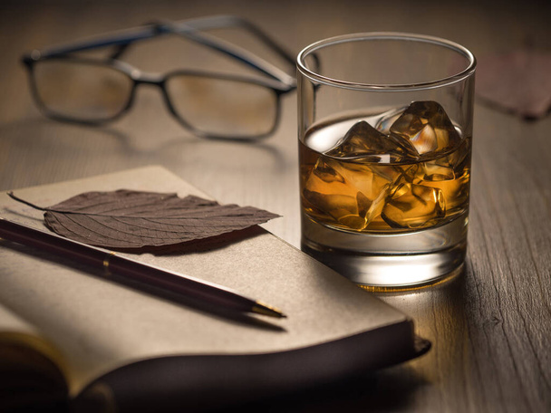 Backlit glass of whisky on the rocks on a wooden table, with notebook, pen and reading glasses - Foto, Imagem
