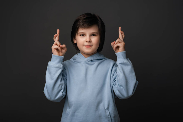 Obrázek roztomilého chlapce 10-12 let v neformálním oblečení držící palce pro štěstí. Lidské emoce, pojetí výrazu obličeje. Studio záběr, šedé pozadí - Fotografie, Obrázek