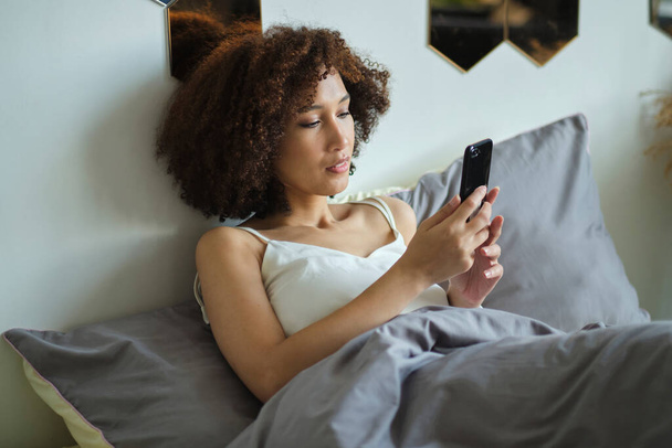 Calm beautiful african woman in pajamas using smartphone while lying in bed isolated. - Foto, Imagem