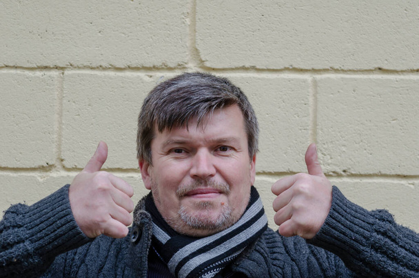 Portret van een volwassen man met twee handen tegen een lichte muur. Een man van middelbare leeftijd houdt zijn duimen omhoog. Lichaamstaal. - Foto, afbeelding