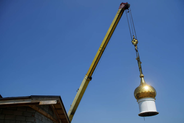 Żuraw podniósł jasną złotą kopułę kościoła chrześcijańskiego do montażu i montażu na dachu budowanej świątyni. Błękitne niebo. Religia i symbole - Zdjęcie, obraz