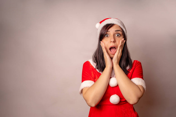 Woman in Santa Claus suit opens mouth and clutches her cheeks in surprise on grey background. Close up of female in Christmas hat in state of shock from amazement. Concept of Christmas celebration - Фото, зображення