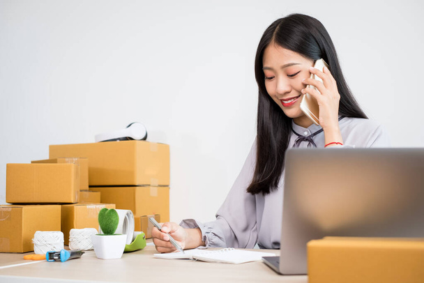 Young Asian woman business owner with many parcel boxes on the table happy online sales job, use your smartphone, get an order from customers, take notes, and make arrangements for delivery by post. - Foto, imagen