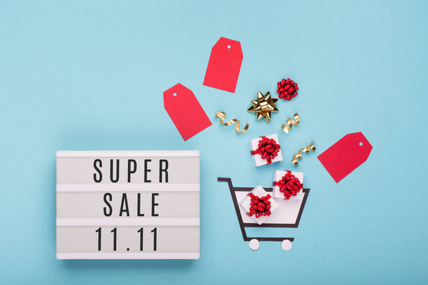 11.11 Super Sale szöveg fehér fénydoboz, bevásárlókocsi, ajándékdobozok és piros címkék kék kék kék papír háttér. Egyedülállók napi koncepció. Online vásárlás Kína. Felülnézet, lapos fekvés, másolás. - Fotó, kép
