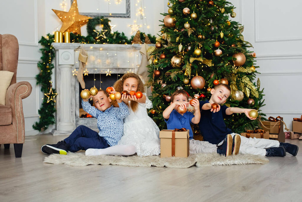 A group of children under a Christmas tree at home. Boys and girls in fancy costumes celebrate winter holidays. Children open gifts by the fireplace. - Fotografie, Obrázek