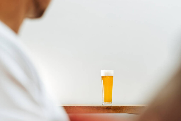 Glass of beer on wooden table with male in foreground, focus on the glass. - Fotografie, Obrázek