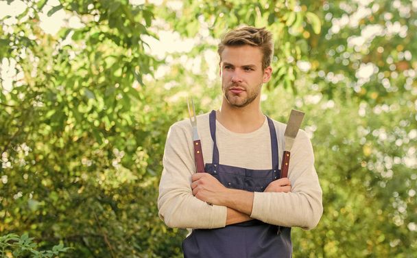 Picnic concept. Bbq chef. Summer weekend. Tools for roasting meat outdoors. Barbeque party. Handsome guy cooking food. Grilling food. Barbecue utensils. Cooking burgers. Man hold barbeque equipment - Valokuva, kuva
