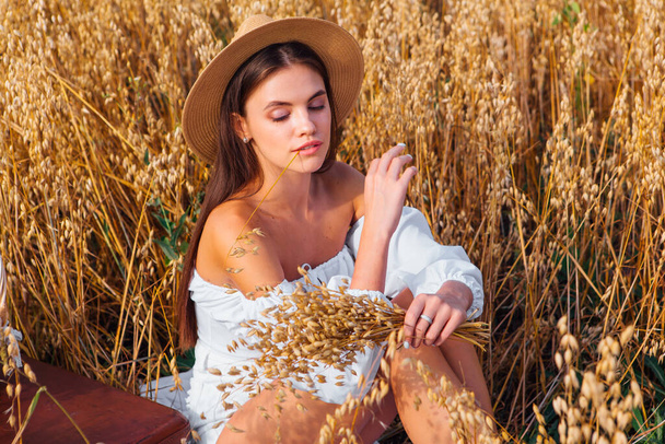 Rural Countryside Scene. Young beautiful woman with long hair dressed in white blouse and straw hat sitting at golden oat field near the basket with ears of oats. - Fotoğraf, Görsel