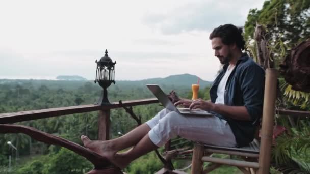 Mladý pohledný muž s vousy pracuje na notebooku v kavárně s výhledem na neuvěřitelný západ slunce. Na volné noze s láskou k cestování, sedí v kavárně a popíjí ovocný koktejl. - Záběry, video