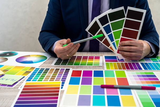 graphic or interior young designer in suit choosing color from swatch sample or catalogue palette guide at workplace. graphic designer with paint color palette samples at table, close up - Photo, Image