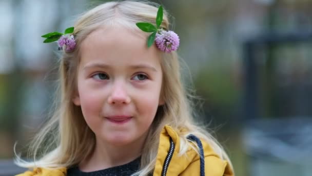 Πορτρέτο ενός υπέροχου κοριτσιού, τριφύλλι λουλούδι στα μαλλιά. χαριτωμένο κορίτσι 5 ετών πονηρά μάτια κοιτάζοντας μακριά με ένα χαμόγελο ματιά στην κάμερα. Κλείστε Shot βίντεο. Πλάνα αργής κίνησης - Πλάνα, βίντεο