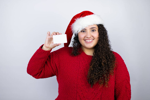 Giovane bella donna che indossa un cappello di Babbo Natale su sfondo bianco sorridente e in possesso di carta bianca - Foto, immagini