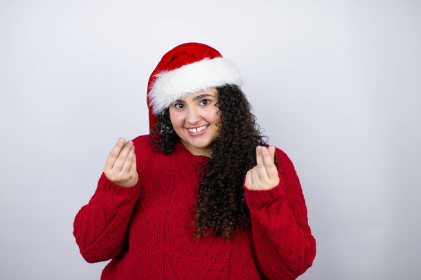 Junge schöne Frau mit Weihnachtsmütze über weißem Hintergrund macht Geld-Geste mit den Händen, verlangt Gehaltszahlung, Millionärsgeschäft - Foto, Bild