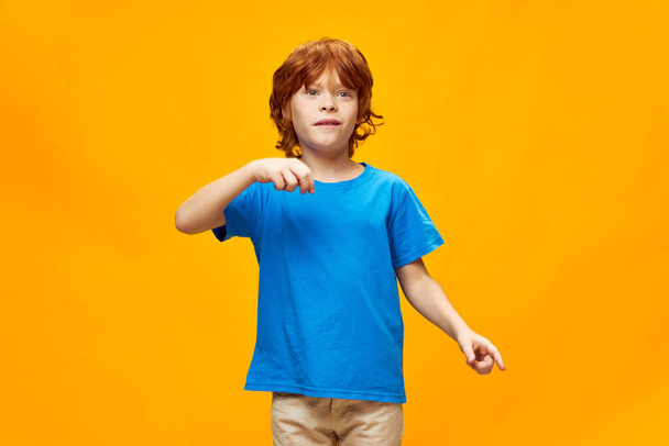 Рыжий ребенок в синей футболке жесты с эмоциями рук желтый изолированный фон  - Фото, изображение