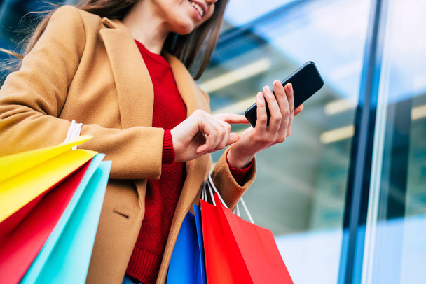 Beautiful trendy young woman with many colorful shopping bags in good mood with smart phone and credit card while walking in the mall during black friday - Fotoğraf, Görsel