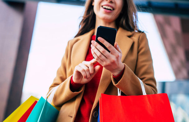 Beautiful trendy young woman with many colorful shopping bags in good mood with smart phone and credit card while walking in the mall during black friday - Φωτογραφία, εικόνα