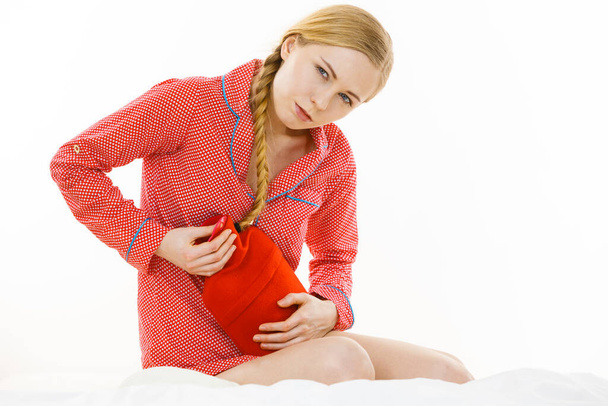 Painful periods and menstrual cramp problems concept. Woman having stomach cramps lying on bed holding hot water bottle feeling very unwell. - 写真・画像
