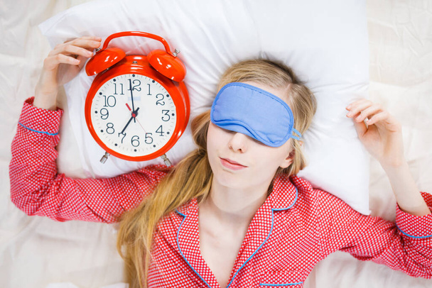 Sleeping young woman wearing cute pink pajamas holding big red old fashioned clock showing sleep time. - Photo, Image