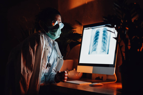 Σύγχρονος ιατρός με υπολογιστή και μάσκα σε σκοτεινό δωμάτιο - Φωτογραφία, εικόνα