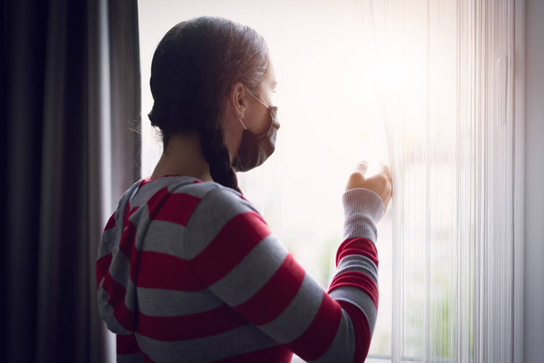 Jovem branca adolescente vestindo máscara protetora, em casa de repouso, olhando para fora da janela com tristeza nos olhos, auto-isolamento devido à pandemia global de Coronavirus COVID-19 - Foto, Imagem