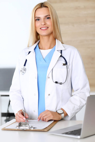 Doctor woman at work in hospital excited and happy of her profession. Blonde physician controls medication history records and exam results. Medicine and healthcare concept - Фото, изображение