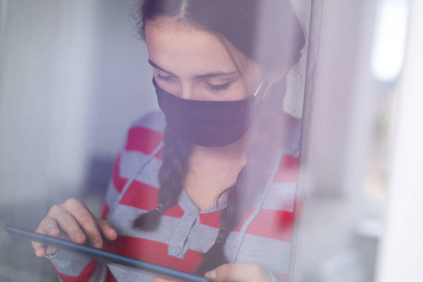 Νεαρό καυκάσιο έφηβο κορίτσι που φοράει προστατευτική μάσκα προσώπου, σε γηροκομείο, κοιτάζει έξω από το παράθυρο με θλίψη στα μάτια της, αυτο-απομόνωση λόγω της παγκόσμιας πανδημίας του COVID-19 Coronavirus - Φωτογραφία, εικόνα