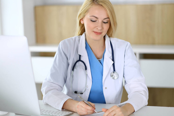 Doctor woman at work while sitting at the desk in hospital or clinic. Blonde cheerful physician filling up medical records form. Data and best service in medicine and healthcare - Photo, Image