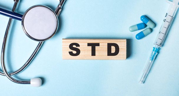 Op een blauwe achtergrond, een stethoscoop, een spuit en pillen en een houten blok met het woord STD. Medisch concept - Foto, afbeelding