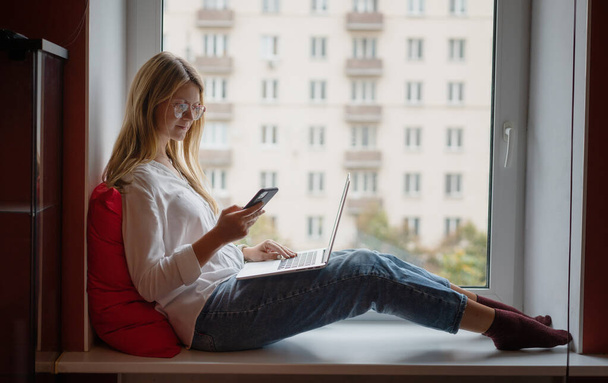 Young business woman with long red hair wearing glasses sitting on windowsill at home using smartphone and laptop - Foto, Imagen